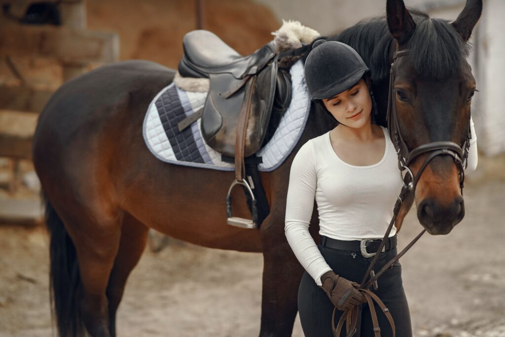 A young woman in equestrian gear stands with her brown horse outdoors, showcasing a bond of trust and care.