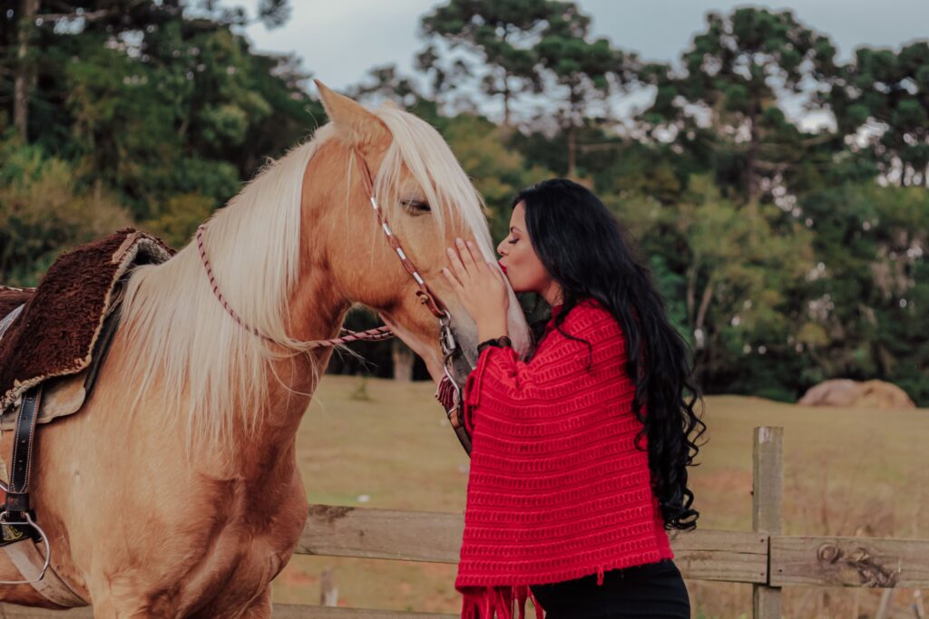 Side view of young female equestrian with long dark hair kissing purebred horse with bridle while spending time together in countryside