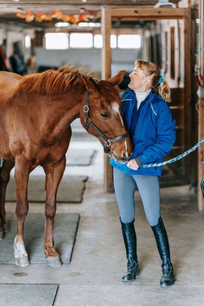 Smiling woman in blue jacket with horse in indoor stable, showcasing bond.